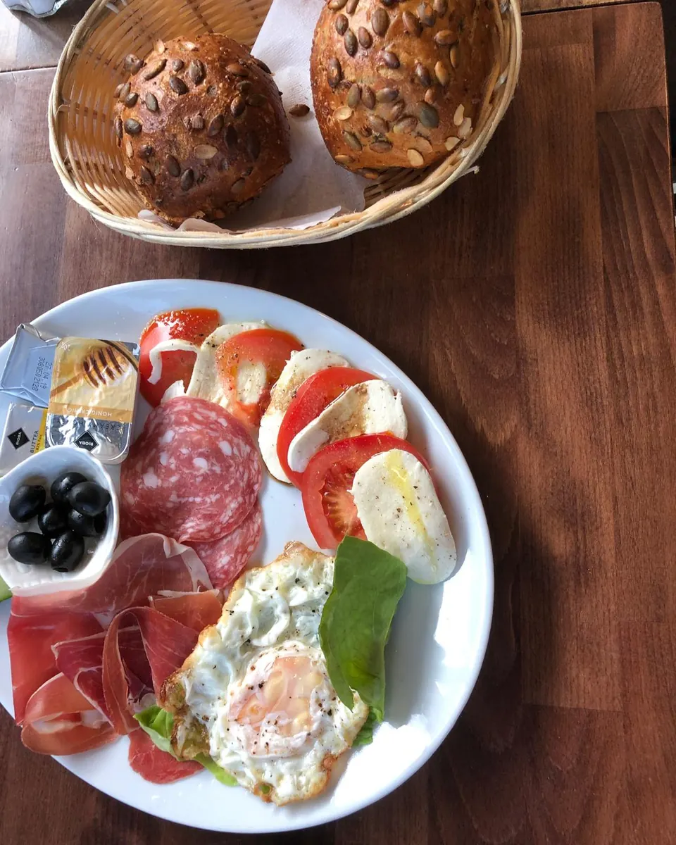 Frühstücksteller mit Spiegelei, Tomate-Mozzarella, Salami, Parmaschinken und Oliven, daneben ein Korb mit Körnerbrötchen