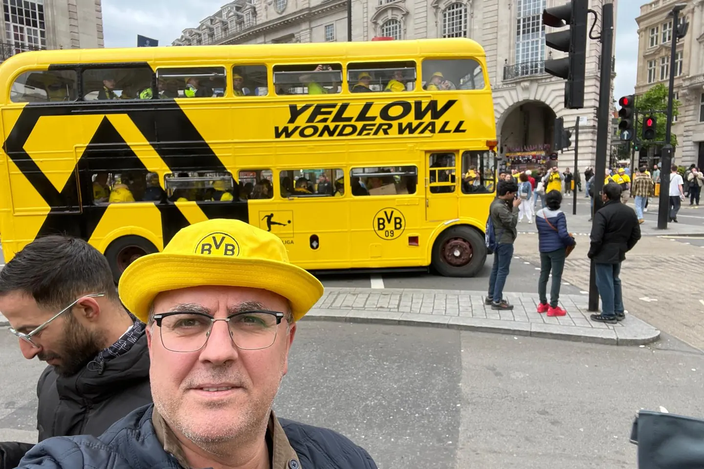 Gelber Londoner Doppeldeckerbus voller Fans, davor ein Fan mit gelbem Hut