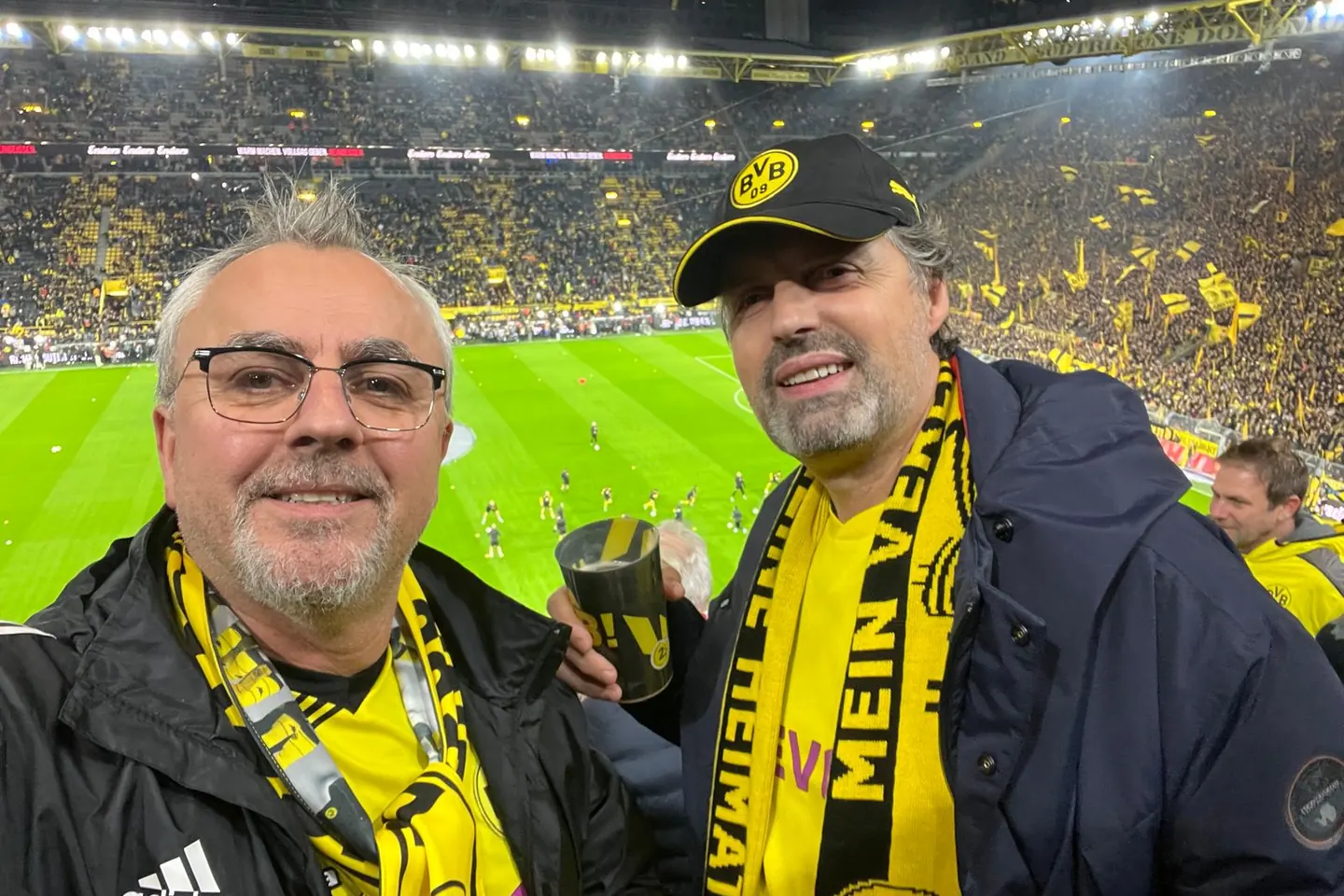 Zwei Fans mit schwarzgelben Schals auf der Tribüne, dahinter die besetzten Ränge