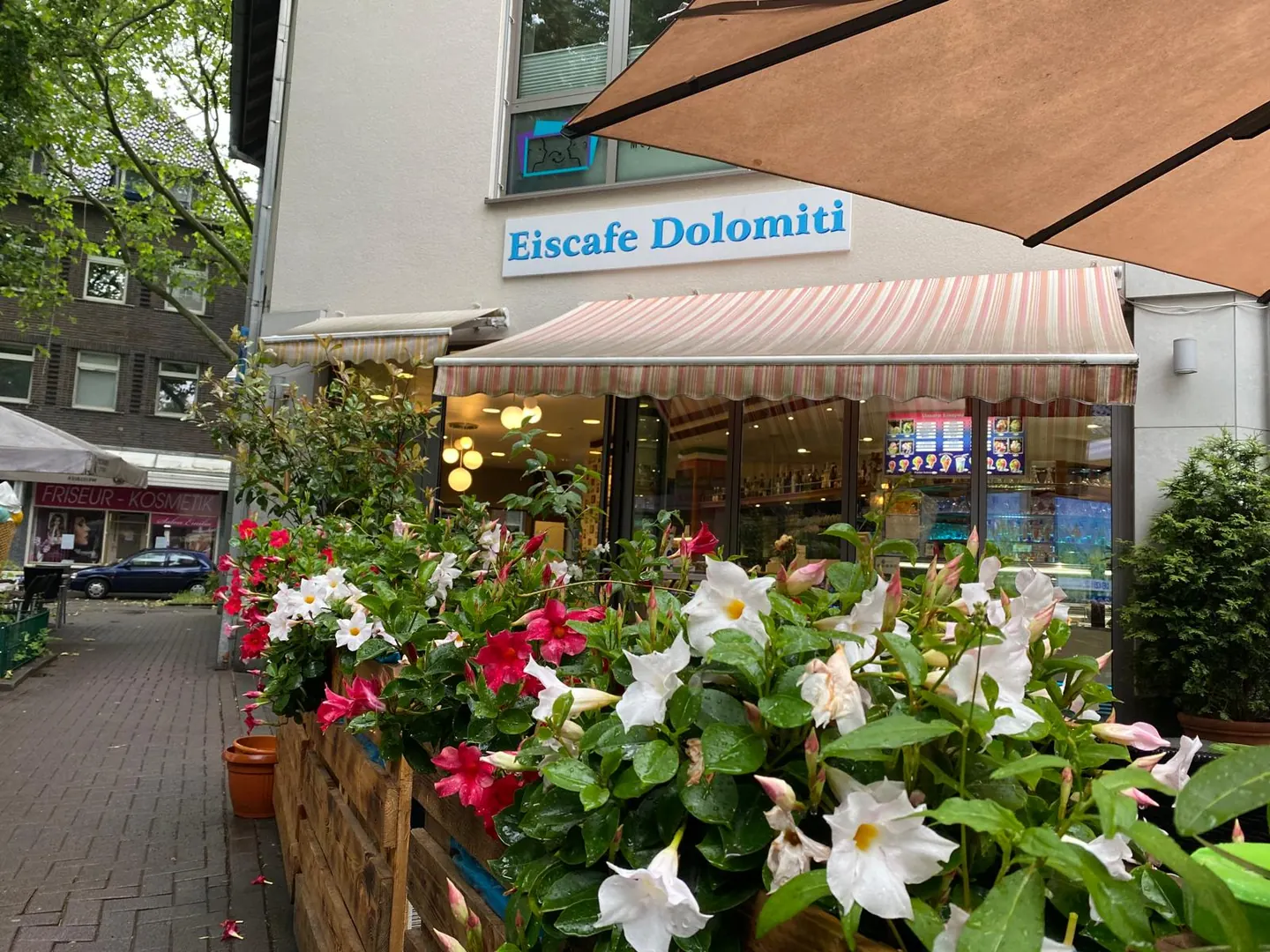 Eingang des Eiscafé Dolomiti unter der Markise, davor Holzkästen mit weißen und roten Blüten