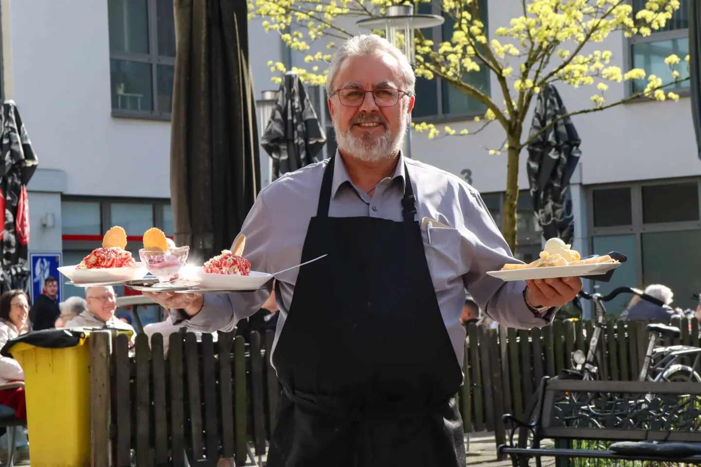 Gastgeber trägt zwei Tabletts mit Eisbechern und Waffeln über die sonnige Terrasse