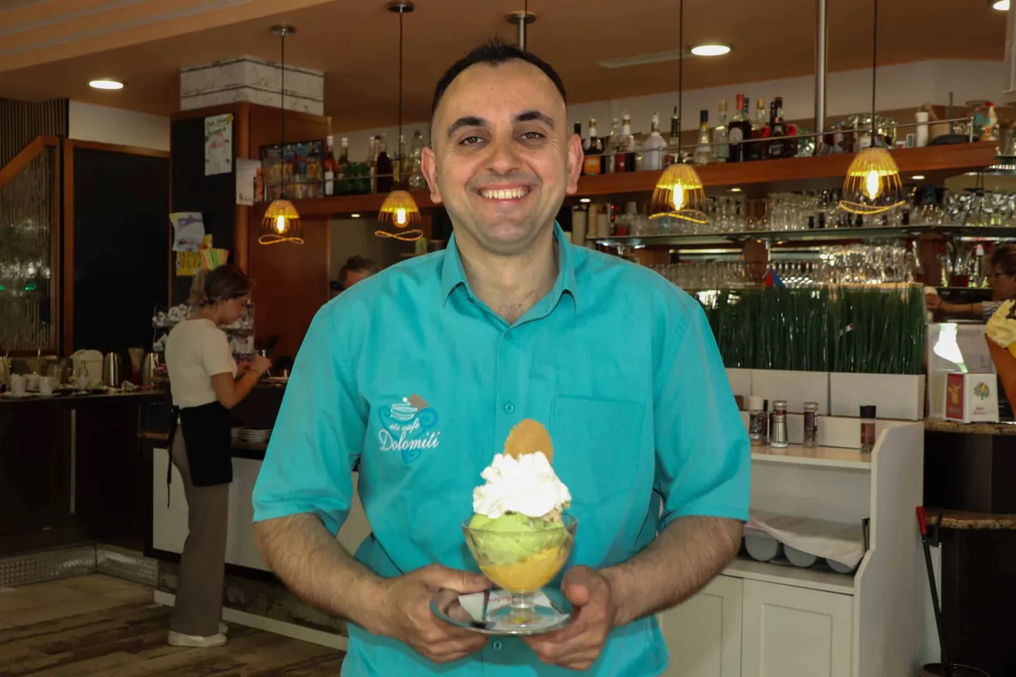 Mitarbeiter im türkisen Dolomiti-Hemd hält einen Eisbecher mit Sahne und Waffel
